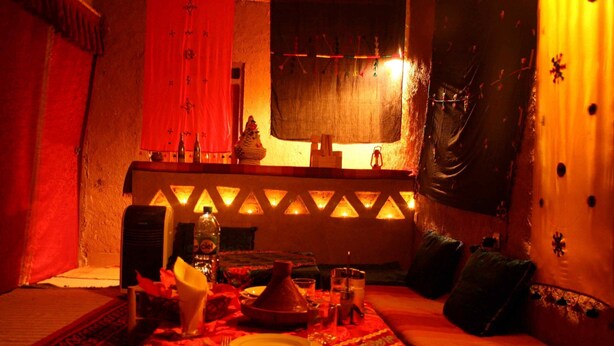 A dimly lit room with a traditional Moroccan-style table set for dining, a sofa, and a decorative wall with a window. - Kasbah Casa Khamlia (Taouz)