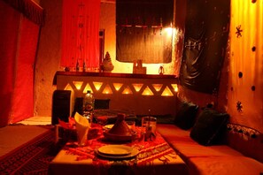 A dimly lit room with a traditional Moroccan-style table set for dining, a sofa, and a decorative wall with a window. - Kasbah Casa Khamlia (Taouz)