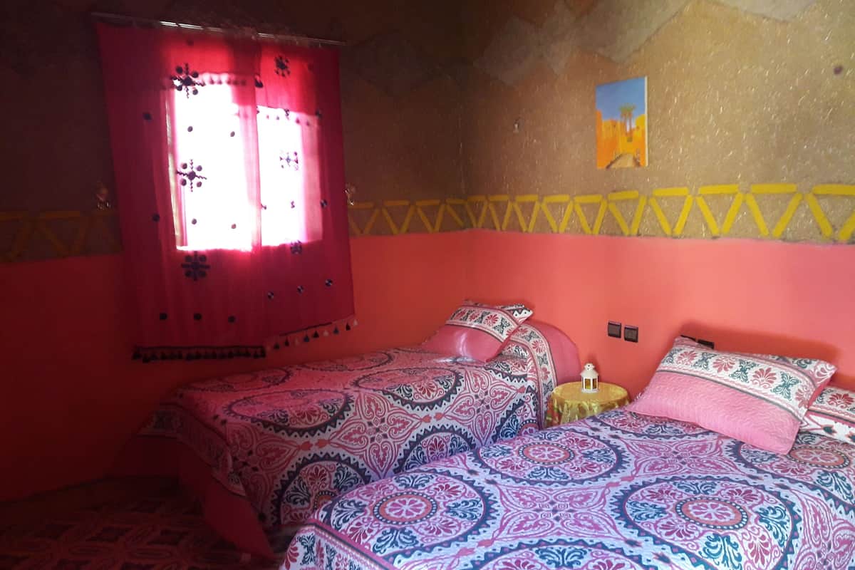 Una habitación con dos camas, una ventana con cortinas rojas, una mesita con un farol y una pared con un motivo geométrico amarillo.