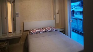 A bedroom with a bed, a ceiling fan, a window with a view of buildings, and a door. - Residencial Atalanta (Bombinhas)