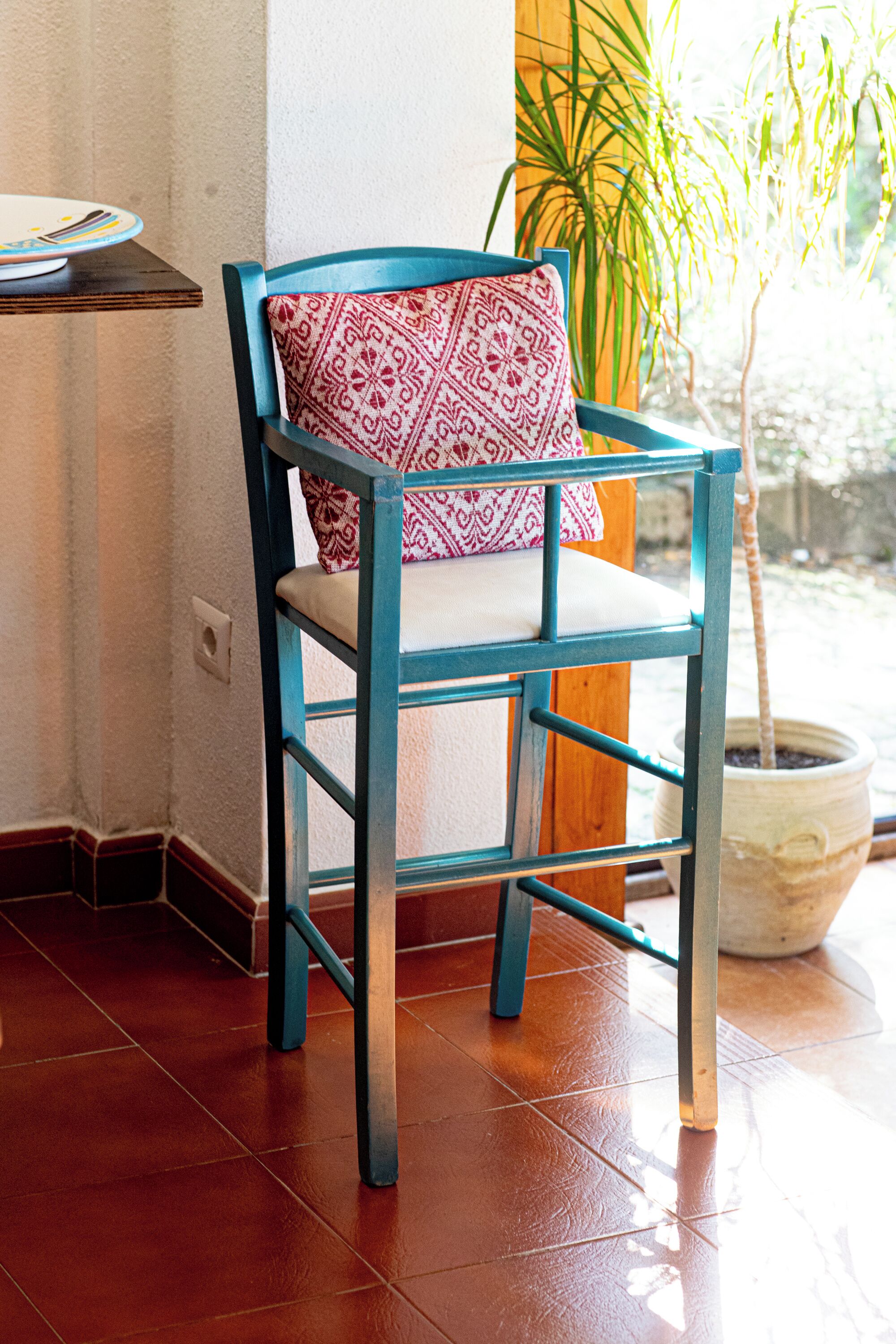 Una silla color turquesa con un cojín estampado en una habitación con suelo de baldosas y una planta en maceta.