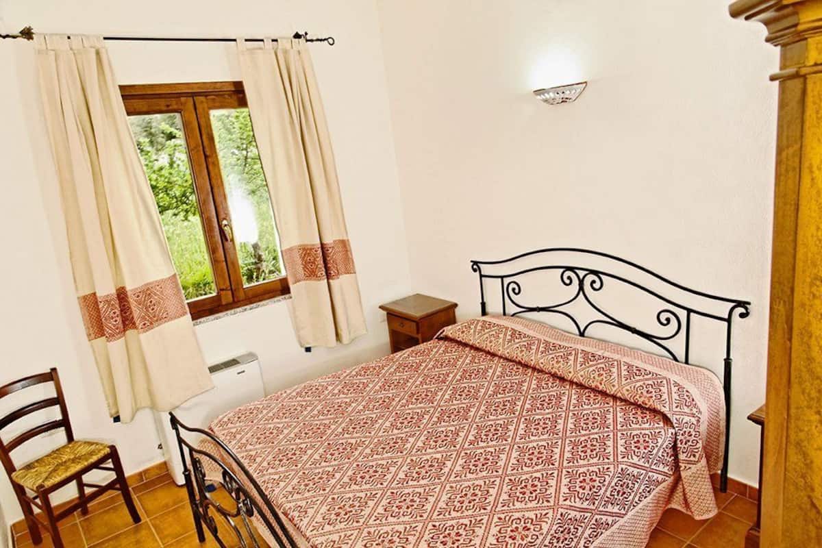 Ein Schlafzimmer mit Bett, Sessel, Nachttisch, Fenster mit Vorhängen und einer Wandleuchte.
