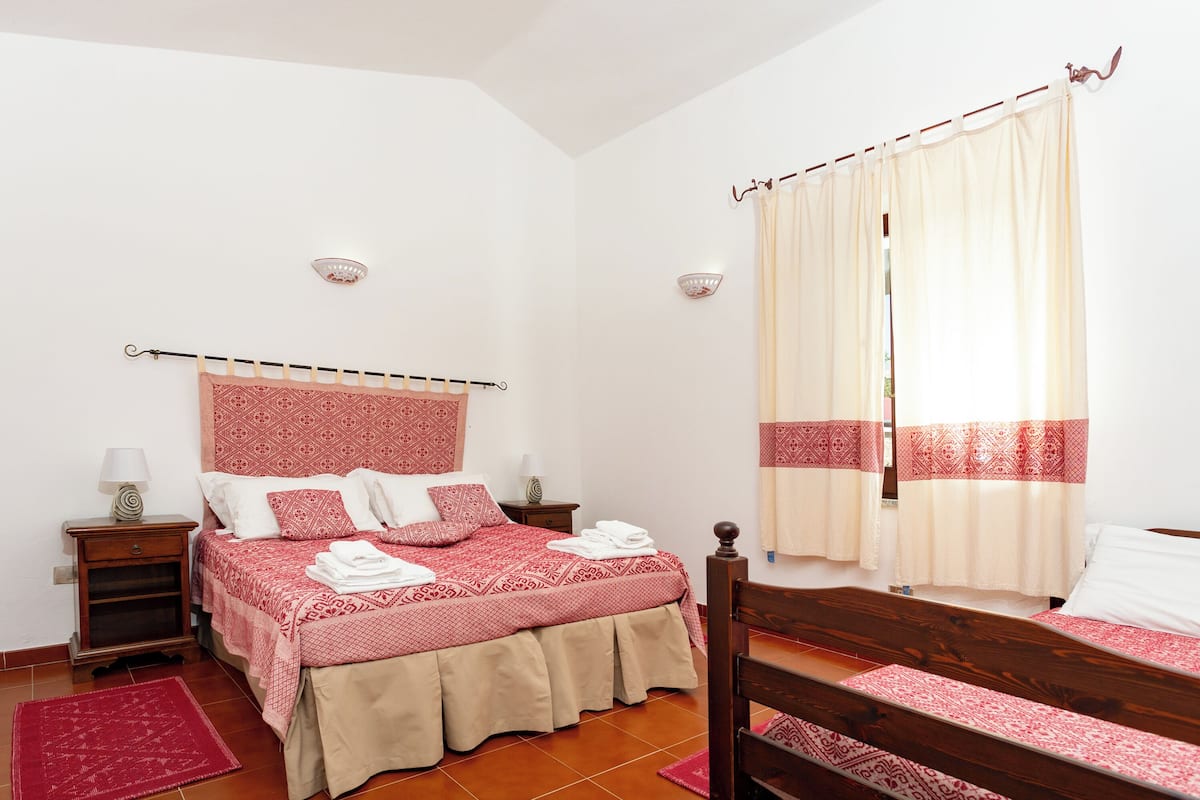 Ein Schlafzimmer mit Bett, Nachttisch, Holzstuhl, einer Bettdecke mit rotem und weißen Muster, einem roten Teppich und einem Fenster mit Vorhängen.
