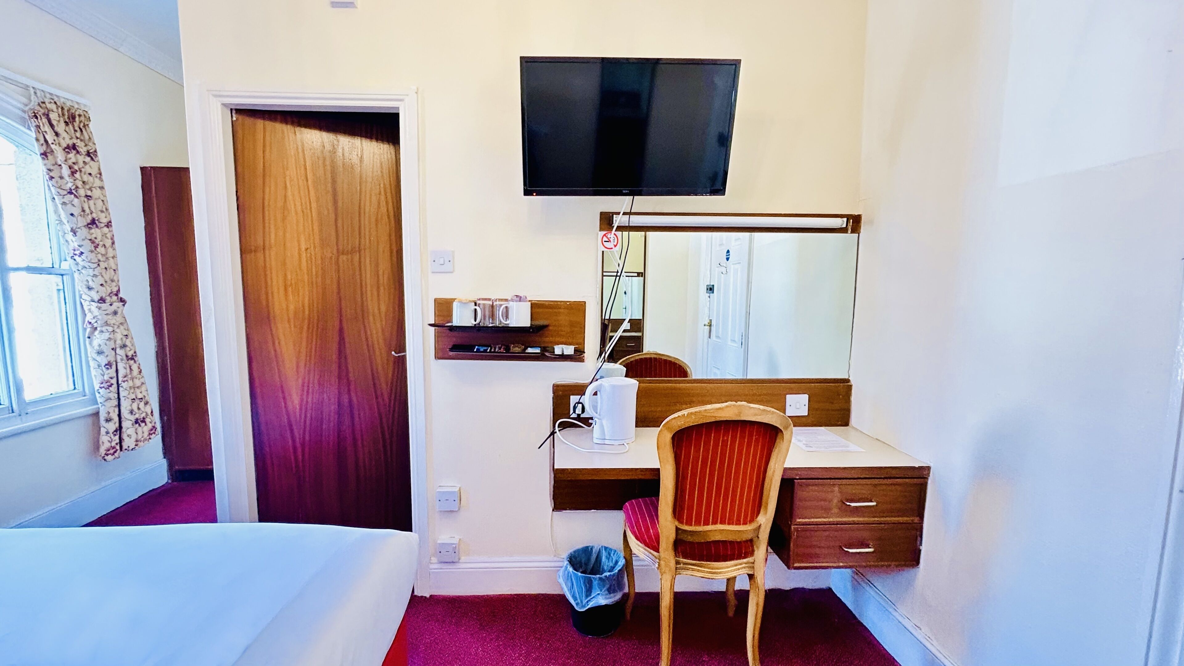 A hotel room with a bed, a desk with a chair, a television, a mirror, a waste bin, and a window with curtains.
