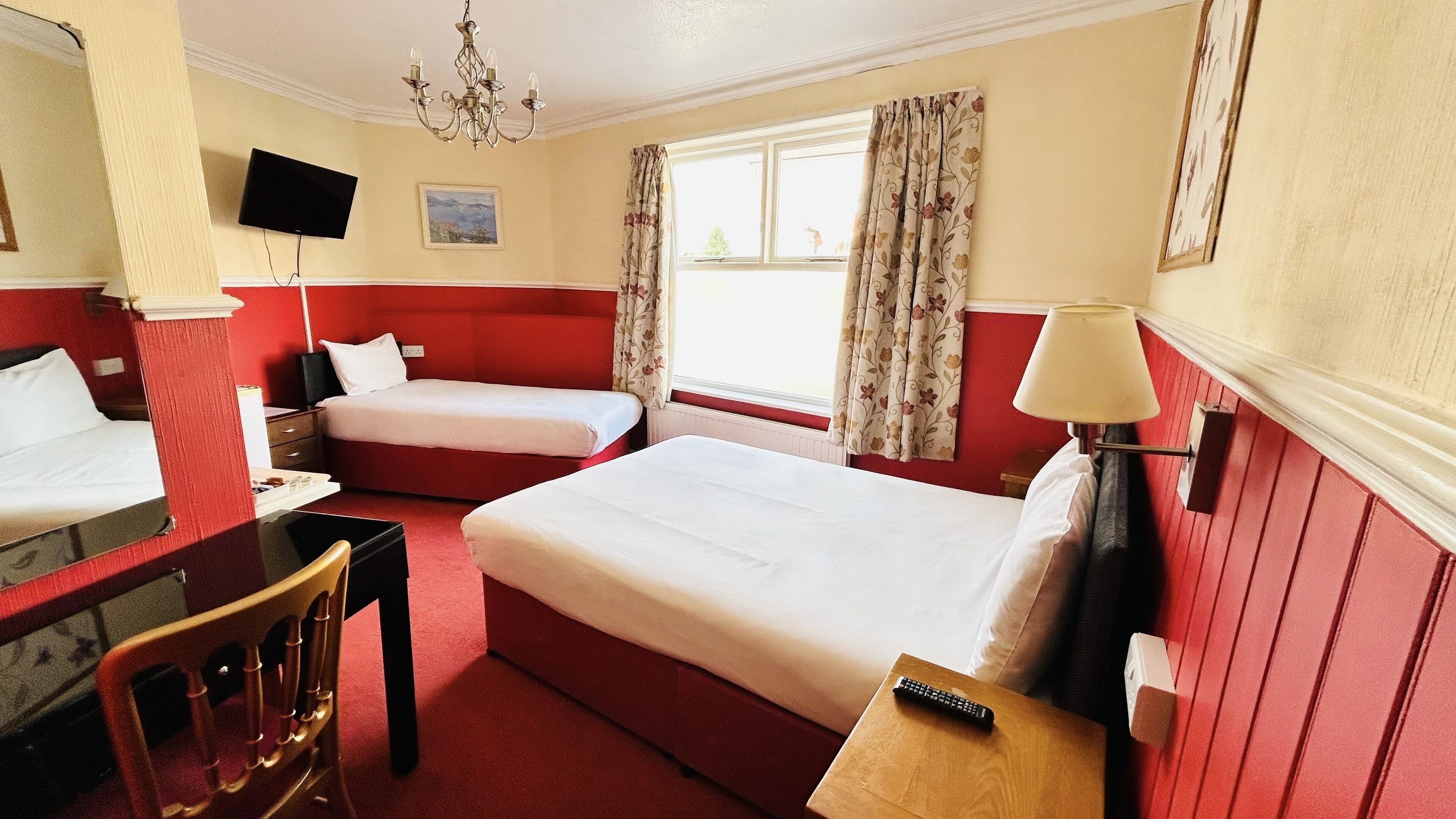 A hotel room with two beds, a desk, a chair, a TV, a window with curtains, and a chandelier.