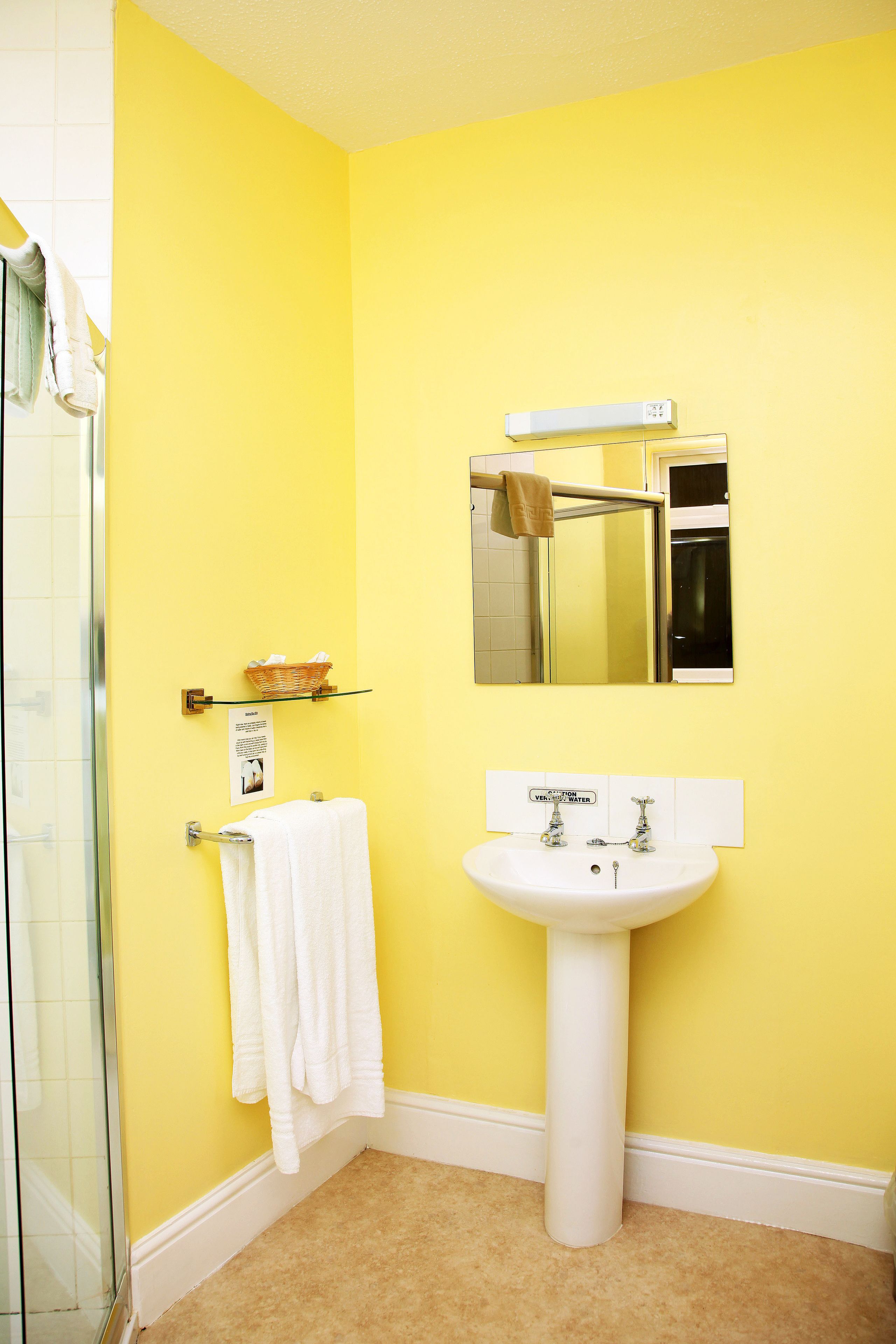 Un baño con pared amarilla, lavamanos blanco y cabina de ducha con mampara de vidrio.