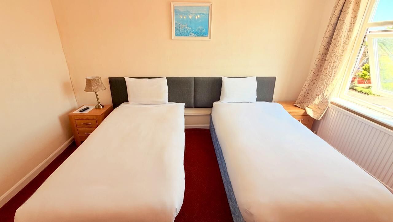 A hotel room with two single beds, a nightstand with a lamp, a framed picture on the wall, and a window with curtains.