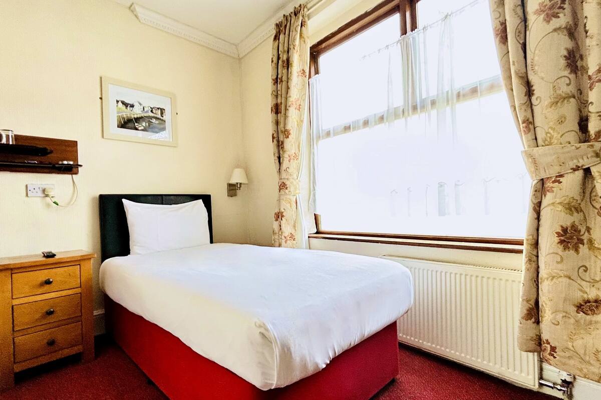 A hotel room with a bed, a nightstand, a lamp, a window with curtains, and a wall-mounted picture.