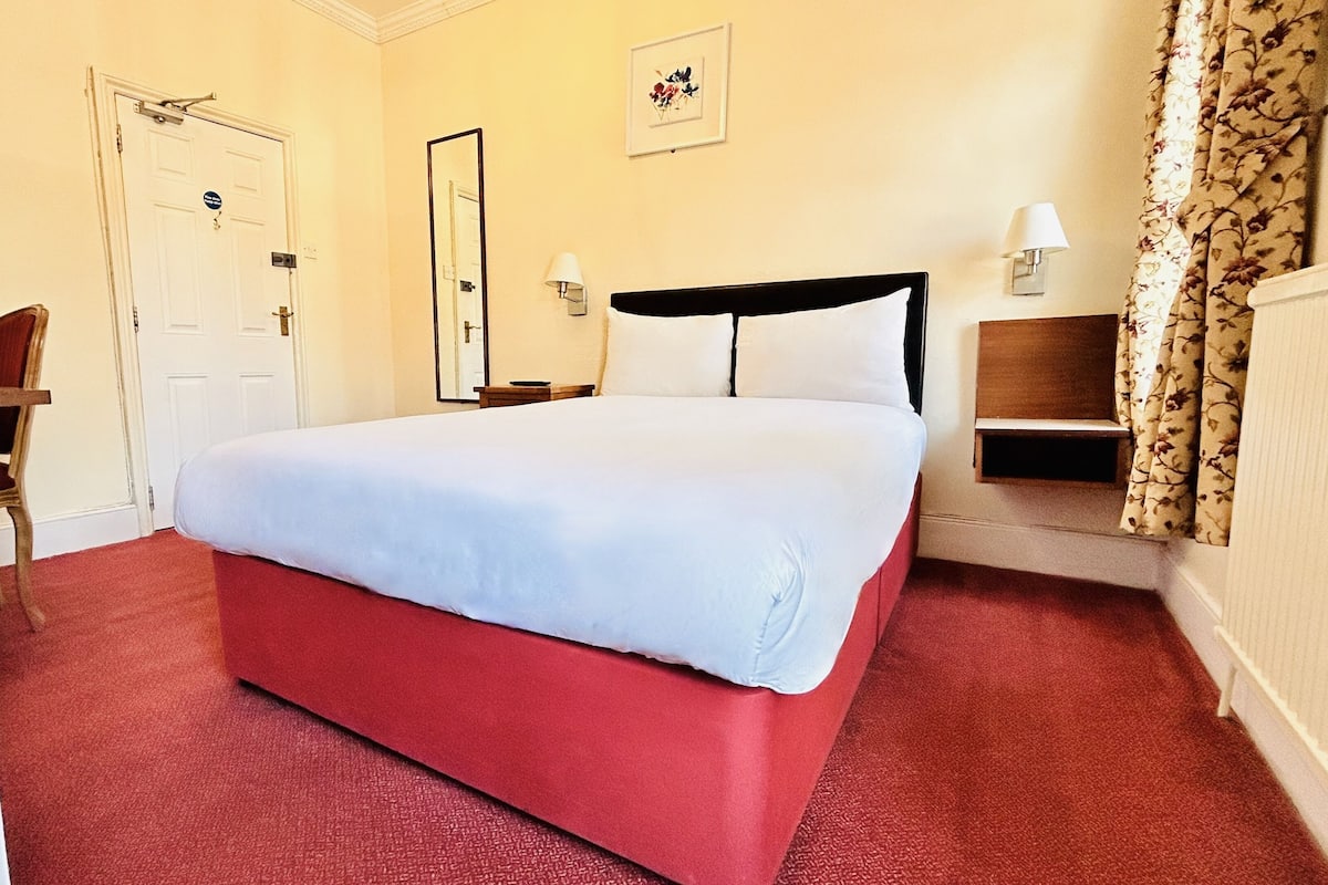 A hotel room with a bed, a chair, a desk, a lamp, a picture on the wall, and a door.