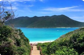 Eine Holztreppe führt zu einem Strand mit klarem türkisblauem Wasser und einem Berg im Hintergrund.