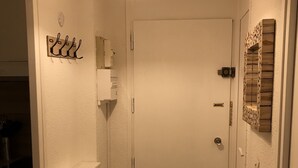 Un pasillo estrecho con una puerta blanca, un estante pequeño, una lámpara de pared y un panel de madera decorativo.