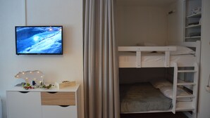 Una cama litera con un televisor montado en la pared, un escritorio y un adorno de oso decorativo, y una cortina entreabierta.