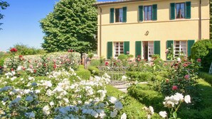 Un bâtiment à deux étages avec des volets verts, entouré d’un jardin bien entretenu où poussent différentes plantes à fleurs et un grand arbre.