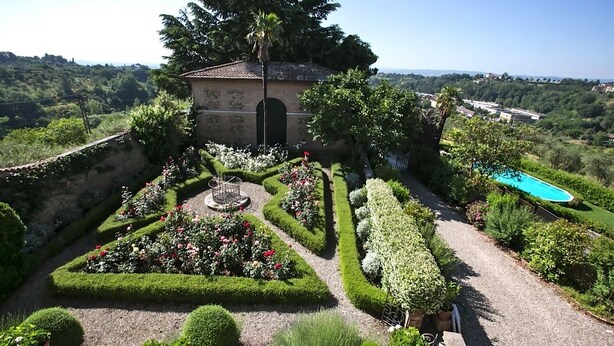 Un jardin aménagé avec une fontaine centrale, une piscine et un petit bâtiment doté d’un toit en tuiles.