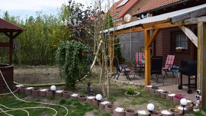 A wooden house with a covered patio, outdoor seating, and a garden with a decorative tree and planters.