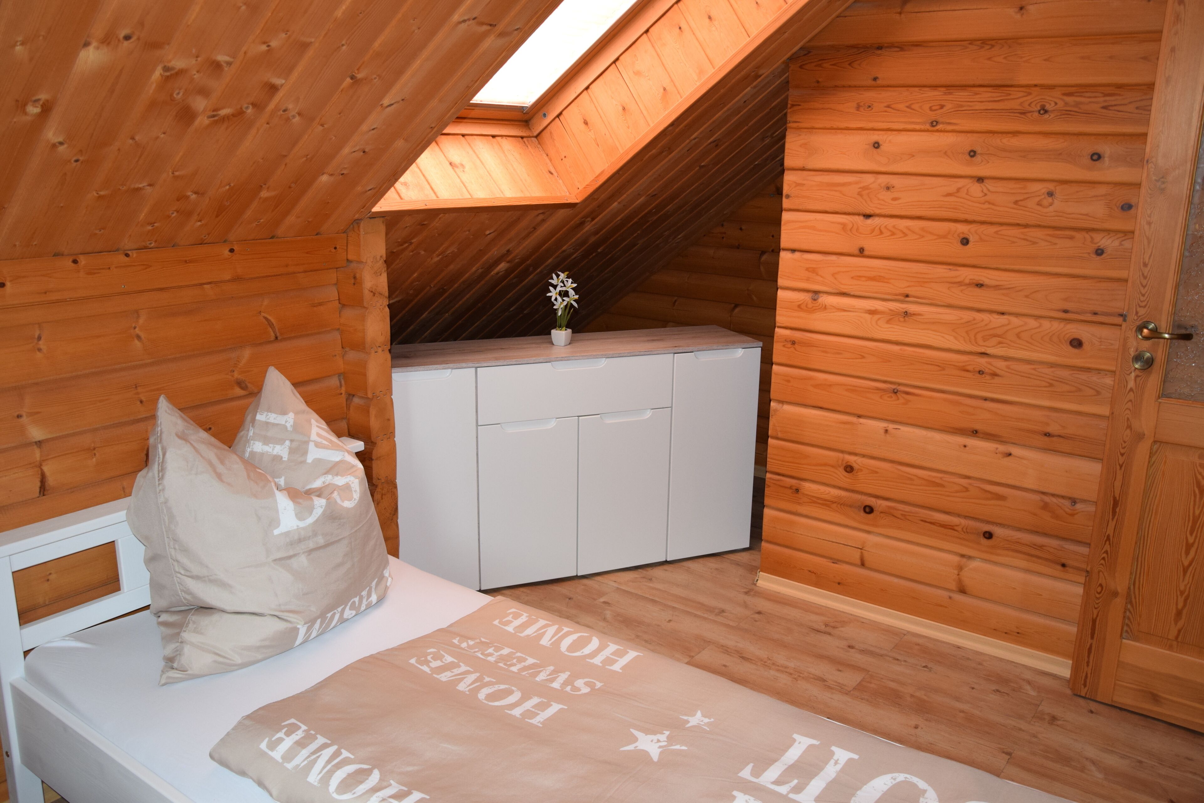 Una habitación pequeña de madera con techo inclinado, una cama con almohada, un armario blanco pequeño y un tragaluz.