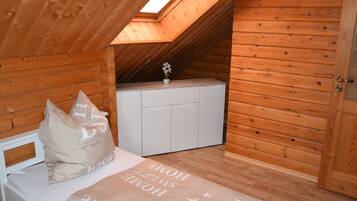Una habitación pequeña de madera con techo inclinado, una cama con almohada, un armario blanco pequeño y un tragaluz.
