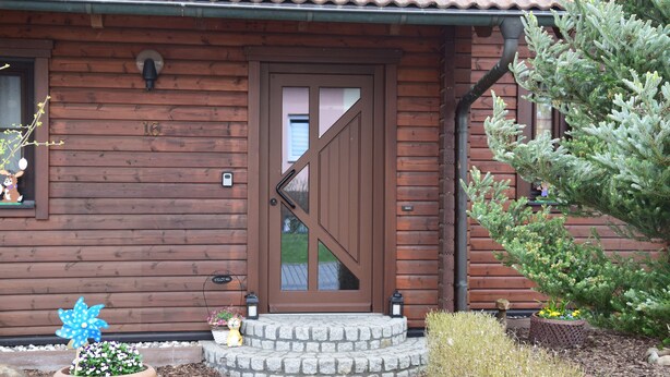 A wooden house with a brown door, a stone staircase, and a potted plant in front.