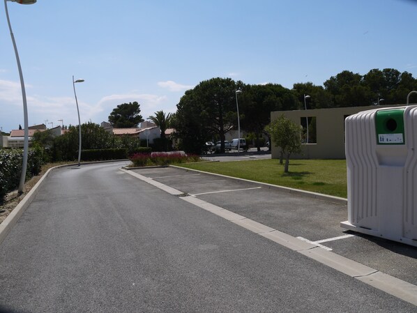 En parkeringsplads med et mobilt toilet, klar himmel og en bygning i baggrunden.