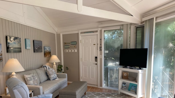 Un soggiorno con un divano grigio, due poltrone, un tavolino da caffè e una televisione su un mobile. Sul pavimento è presente un tappeto decorato, e una porta dà accesso a un balcone con vista sul verde.
