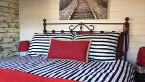 Een bed met een gestreepte sprei en een rood kussen, een schilderij van een spoorlijn aan de muur, en een bakstenen muur links.