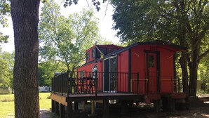 Een rode treinwagon met een balkon en een zwarre reling, omgeven door bomen en een gazon.