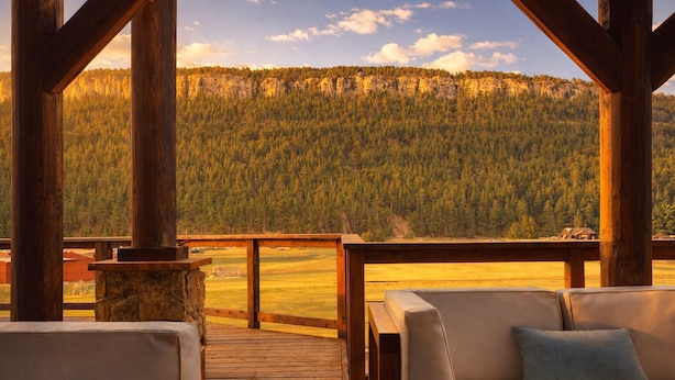 A wooden deck with a sofa and a stone fireplace, overlooking a forested valley.
