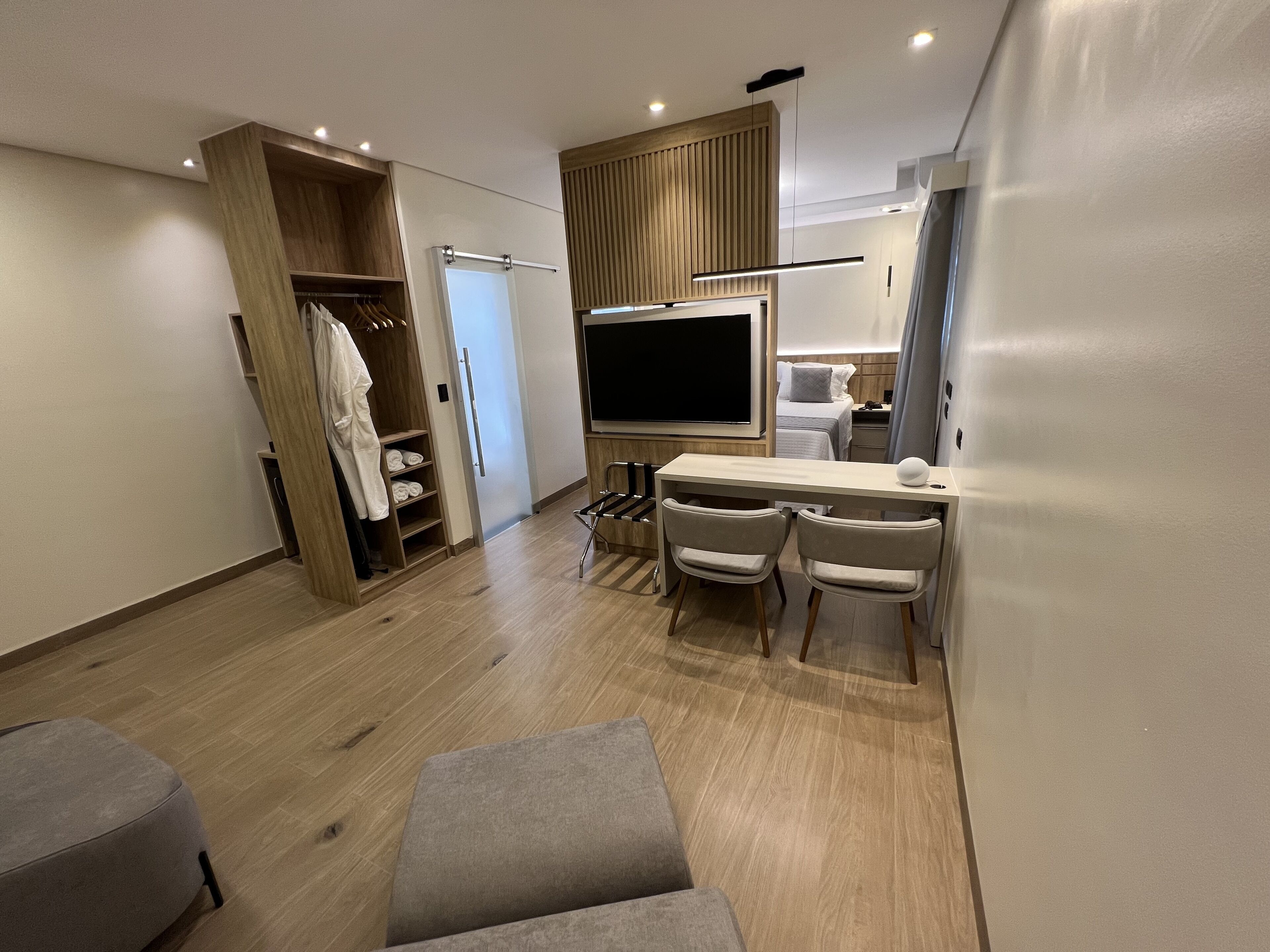 A modern hotel room with a wooden wardrobe, a flat-screen TV, a desk with two chairs, and a small seating area.