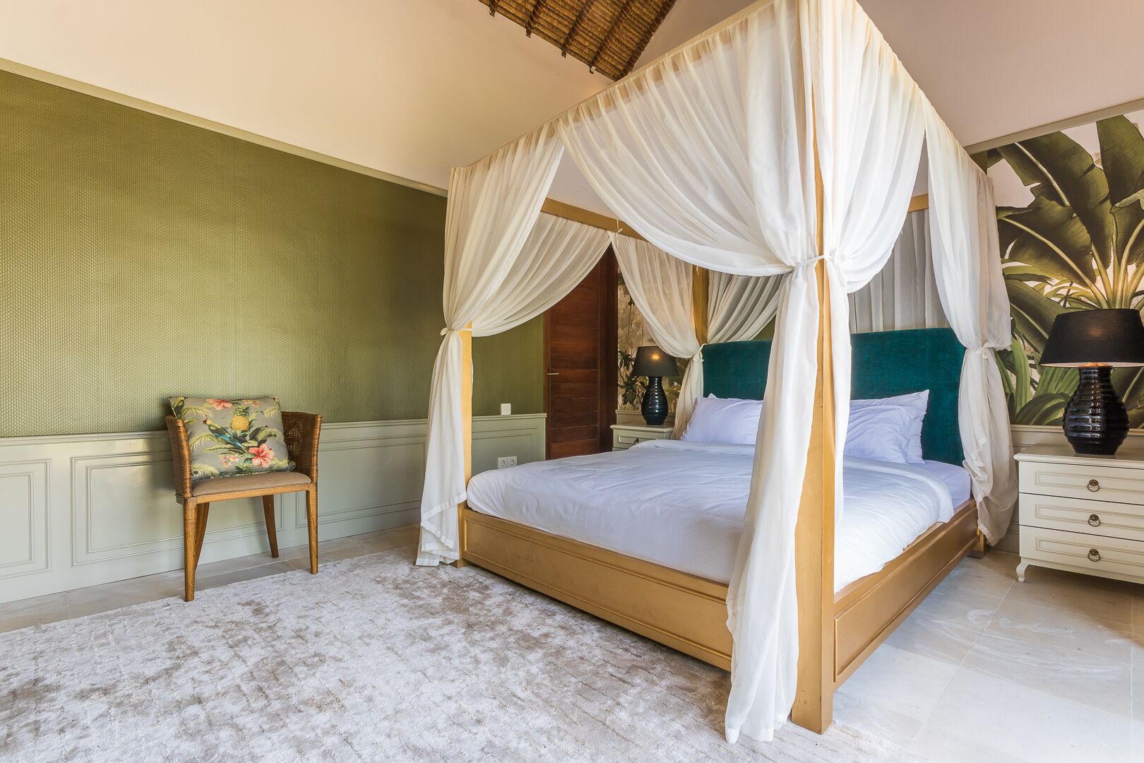 Una cama con dosel y cuatro columnas, ropa de cama blanca, una mesita de noche con lámpara, un sillón con cojín floral y una pared de acento verde.