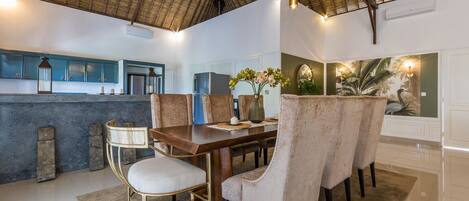 A dining area with a wooden table, cushioned chairs, and a thatched ceiling.