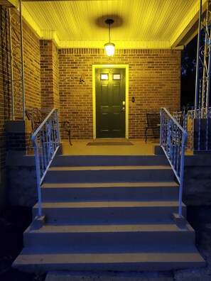 Un conjunto de escalones exteriores que conducen a una puerta, con un accesorio de iluminación encima, rodeados de muros de ladrillo y barandas metálicas.