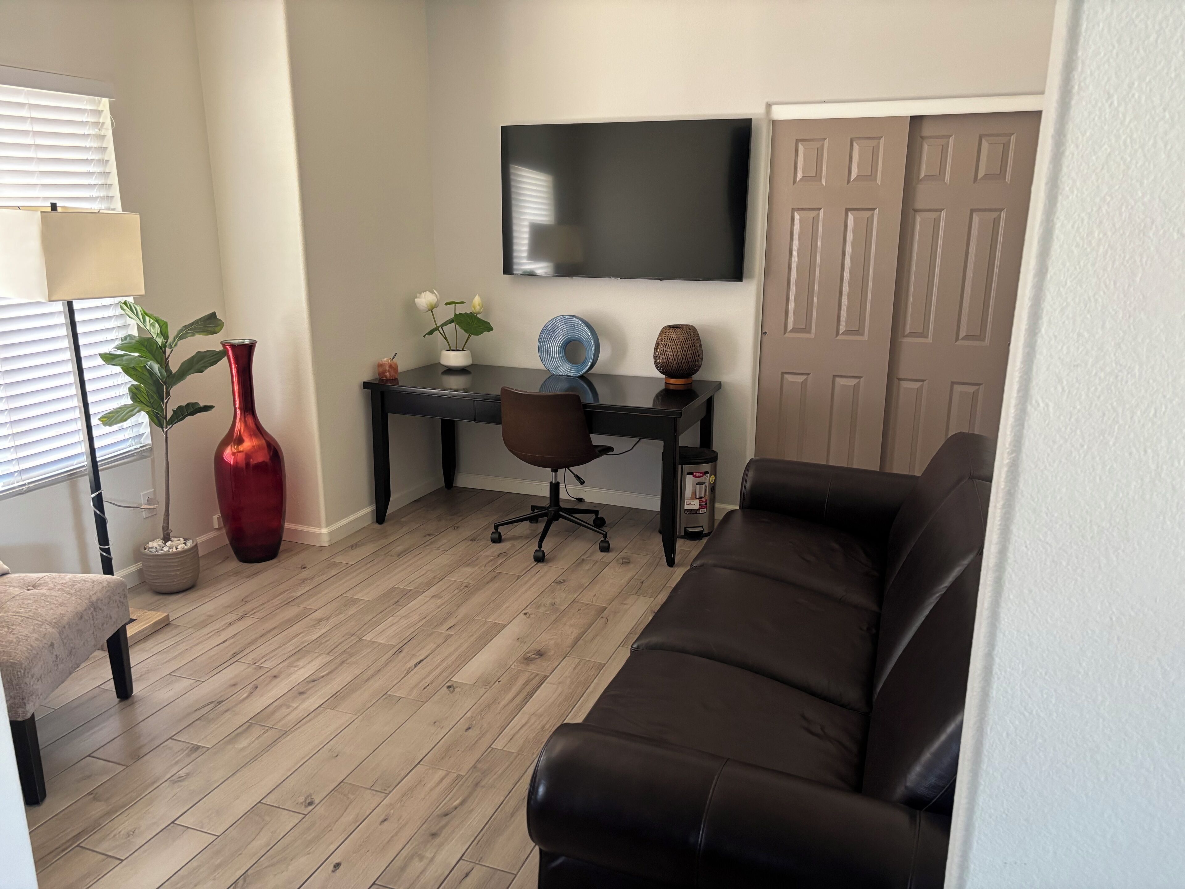 A living room with a black leather sofa, a desk with a chair, a TV mounted on the wall, a potted plant, and a vase.