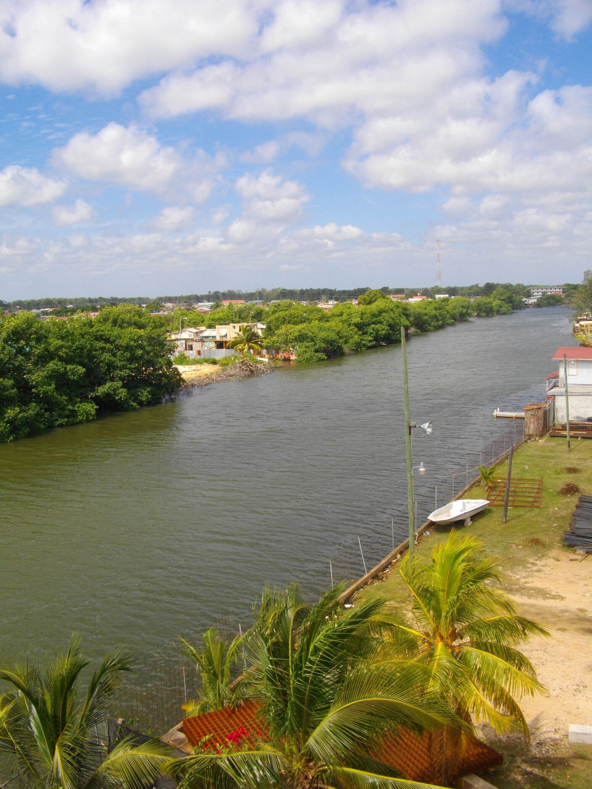 A river with a boat docked on the right, surrounded by greenery and buildings in the background.