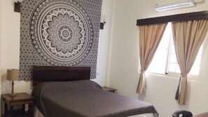 Un dormitorio con cama, mesita de noche, lámpara, tapiz, ventana con cortinas y escritorio.