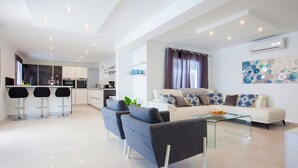 En moderne stue med sofa, stole og et sofabord i glas. Der er et køkkenområde med barstole og hårde hvidevarer.