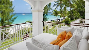 A balcony with a bed, white railing, and a view of the ocean and palm trees. - Condo Old Trees 6 - Firefly (Paynes Bay)