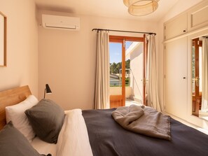 A bedroom with a bed, a nightstand, an air conditioning unit, a window with curtains, and a view of the outdoors.