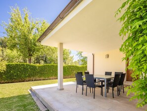 Une terrasse couverte, avec une table et des chaises, entourée d’une haie et d’arbres.