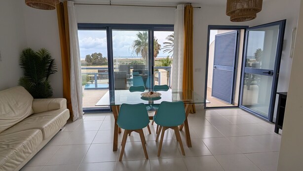 Una sala de estar moderna con una mesa de comedor de vidrio, sillas color turquesa, un sofá color beige y un amplio ventanal corredizo que da a un balcón con vistas a palmeras y edificios.
