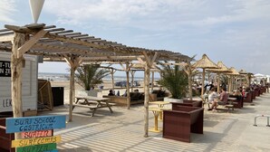 A beachside area with wooden structures, picnic tables, and a sign listing activities like surf school and parties.