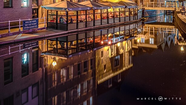 A canal-side building with multiple stories, a restaurant with outdoor seating, and a reflection in the water.