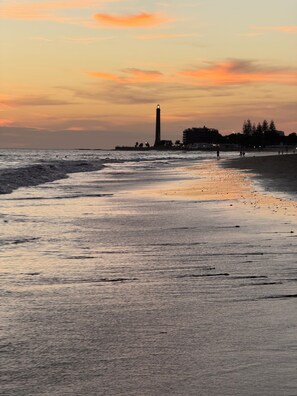 Een strand met een vuurtoren in de verte, golven die de kust naderen, en een heldere lucht.
