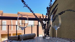 Una mesa metálica redonda con dos copas de vino y un menú enrollado, ubicada en un balcón con vistas despejadas a edificios.