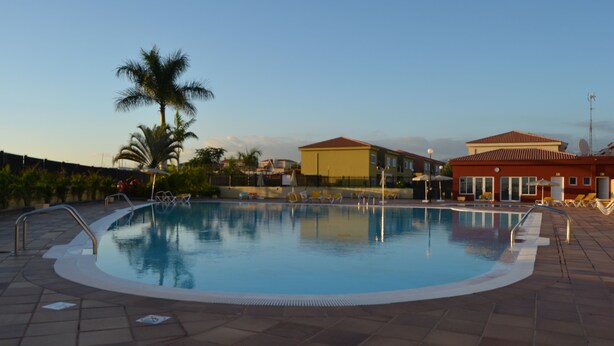 Una piscina con un borde de baldosas, palmeras y edificios al fondo.