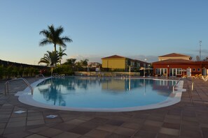 Une piscine avec une terrasse carrelée, des palmiers et des bâtiments en arrière-plan.