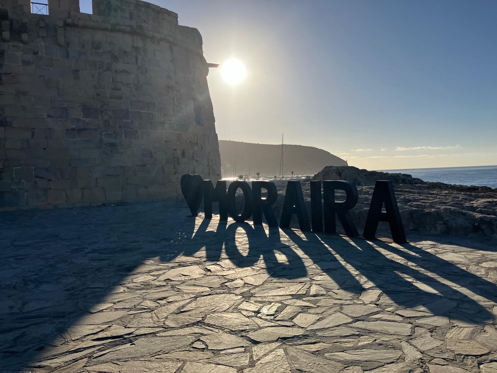 Un camino de piedra con un gran letrero que dice 'MORARA' en el centro, que lleva a una zona costera con un muro de piedra y un cielo despejado.