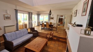 Ein großzügiger Wohnbereich mit einer Couch, Esstisch und Stühlen, einer Lampe und Blick durch große Schiebetüren auf eine Terrasse.