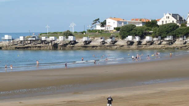 Una spiaggia con persone che nuotano e passeggiano, case sullo sfondo e torri di salvataggio.