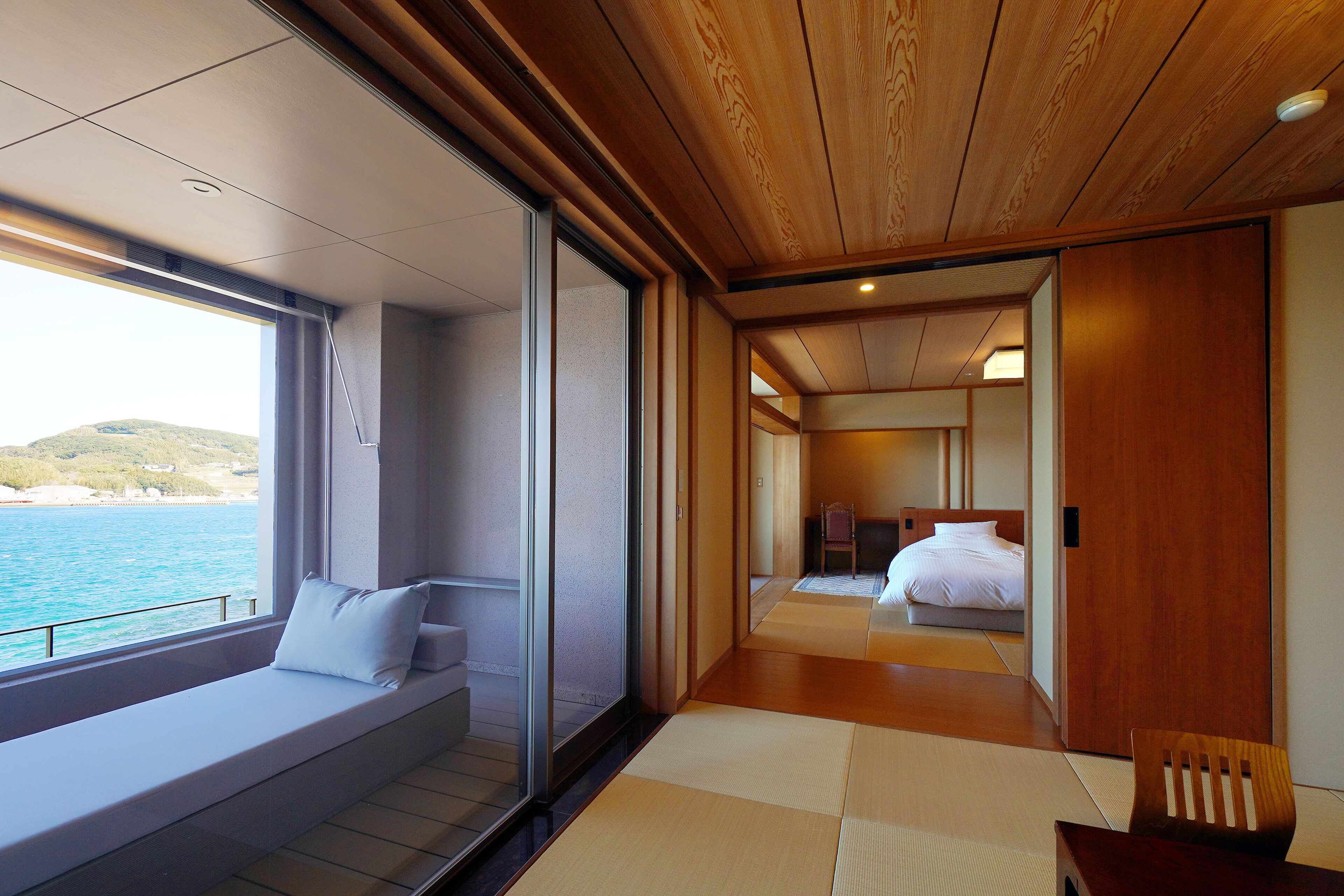 A modern hotel room with a large window overlooking the sea, a wooden ceiling, and a traditional Japanese tatami mat floor.