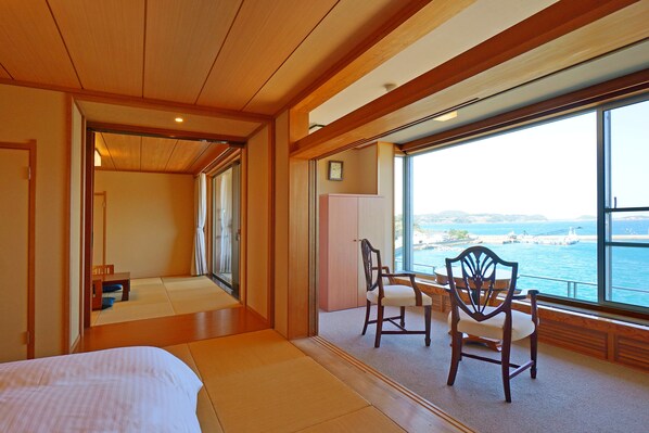 Une chambre avec un lit, deux chaises, une petite table et une vue sur la mer grâce à de grandes fenêtres.